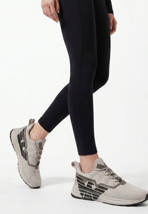 Persoon die beige en zwarte sportsneakers draagt met zwarte legging, met een zijaanzicht van de schoenen en onderbenen.