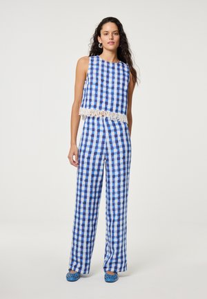 Frau steht und trägt ein blau-weiß kariertes ärmelloses Oberteil mit Fransen am Saum und passende weite Hose mit blauen Schuhen.