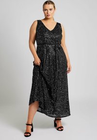 Vestido negro sin mangas con escote en V, cubierto de lentejuelas, con un lazo en la cintura y un dobladillo asimétrico. Combinado con sandalias negras de tacón bloque.