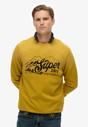 Geel sweatshirt met zwart berggrafiek en tekst "Super Dry Fine Goods Co". Heeft een ribbelhalslijn en lange mouwen. Gedragen over een geruit shirt.