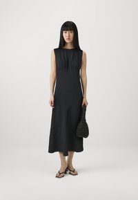 Crna haljina bez rukava s prilagođenim korzetom i lepršavom suknjom, izrađena od glatke tkanine. Manekenka drži teksturiranu crnu torbu i nosi japanke.