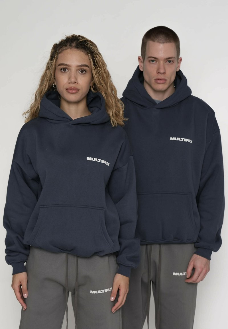 Felpe con cappuccio blu navy con il logo "MULTIPLY" bianco, dotate di tasche a marsupio e vestibilità ampia. Pantaloni della tuta grigi con loghi abbinati.