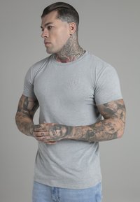 Grijze t-shirt met korte mouwen en een ronde halslijn, voorzien van een subtiel embleem op de borst en een aansluitende snit die getatoeëerde armen toont.