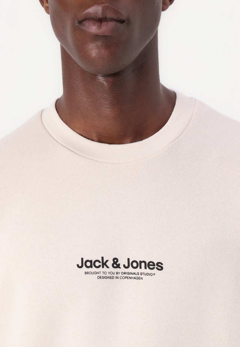 Lichtbeige T-shirt van zachte katoen, met een ronde halslijn en zwart bedrukte logo-details op de borst.