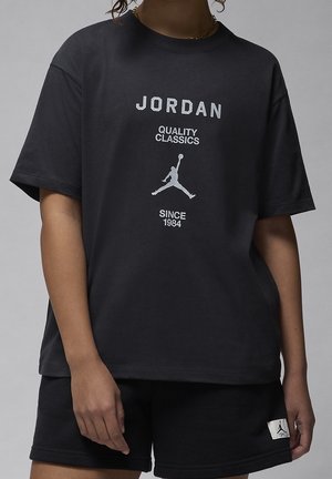 Osoba nosząca czarną koszulkę Jordan z białym logo i tekstem oraz czarne szorty z małą białą naszywką z logo Jordan.