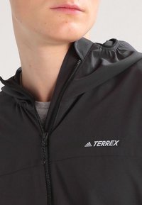 Svart huva jacka gjord av lätt material, med en framdragkedja och "TERREX"-logotyp i vitt på vänster bröst.