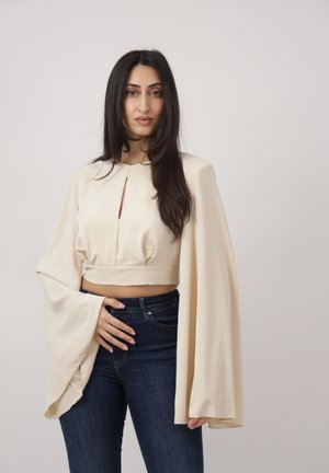 Kobieta o długich ciemnych włosach, ubrana w kremowy, długi top z szerokimi rękawami i ciemnoniebieskie dżinsy, pozująca na jednolitym tle.