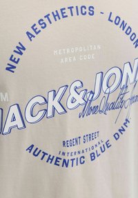 Smėlio spalvos audinys su mėlynu ir baltu tekstu, kuriame matyti "JACK & JONES" bei frazės kaip "Nauja estetika – Londonas", "Regent Street" ir "Autentiškas mėlynas DNM".