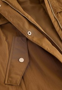 Primer plano de una chaqueta marrón que muestra un botón de presión metálico, cremallera y un bolsillo con solapa con detalles de costura.