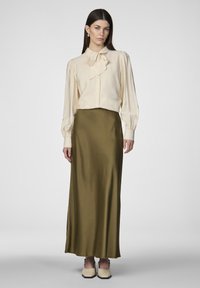 Blouse crème avec un nœud attaché au col, associée à une jupe en satin vert olive élégante. Modèle portant des chaussures de couleur claire.