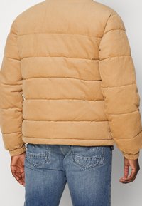 Persona che indossa un giubbotto puffer in corda color sabbia e jeans di denim blu, vista da dietro su uno sfondo semplice.