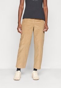 Beige kord pantalone s sproščenim krojem, z stranskimi žepi in teksturirano tkanino. V kombinaciji s svetlimi teniskami in temnim zgornjim delom.