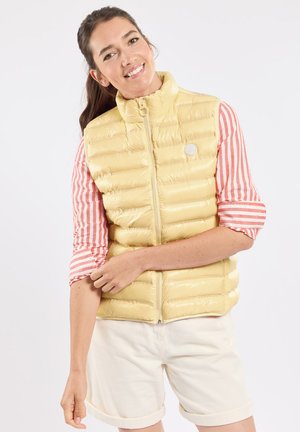 Femme souriante ajustant la manche de sa chemise rayée, portant une veste matelassée sans manches jaune clair et un short clair retroussé, se tenant devant un fond blanc.