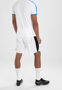 Man som står i vita sportkläder med blå och svarta detaljer, vita strumpor och svarta fotbollsskor, sedd bakifrån mot en enkel bakgrund.