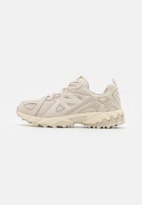 Beige New Balance terepfutó cipő hálós panelek, texturált talp és fűzős záródás jellemzi, a jobb oldalról látható profilban, fehér háttéren.