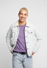 Light blue denim jacket with cream sherpa collar, paired with a purple t-shirt. Classic button-up design and light blue jeans.