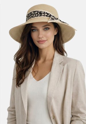 Woman with long brown hair wearing a wide-brim straw hat with a black and white patterned ribbon, beige blazer, and white top against a plain background.