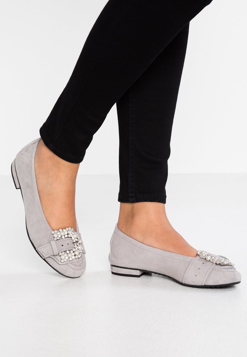 Pieds chaussés de ballerines en daim gris clair ornées de boucles embellies de perles, associées à un pantalon noir ajusté sur un fond blanc uni.