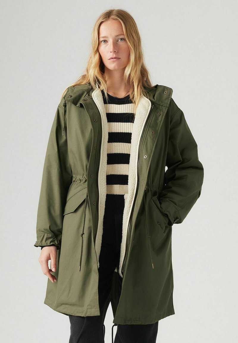 Parka impermeabile verde con cappuccio, vita regolabile con cordino e fodera in finta pelliccia. Presenta grandi tasche frontali e chiusura con zip.