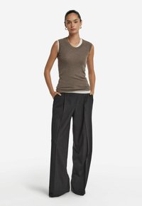 Kobieta z związanymi włosami, nosząca kolczyki w kształcie kół, bezrękawnik w kolorze brązowym na białej koszuli oraz luźne czarne spodnie szerokie w nogawkach, z rękami w kieszeniach.