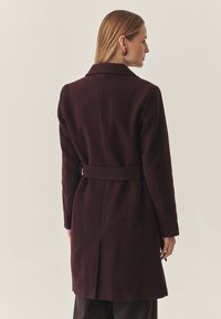 Burgundský vlněný kabát s tvarem na míru, výstřihem do "V", páskem v pase a rozparkem vzadu, který zdůrazňuje hladkou texturu.