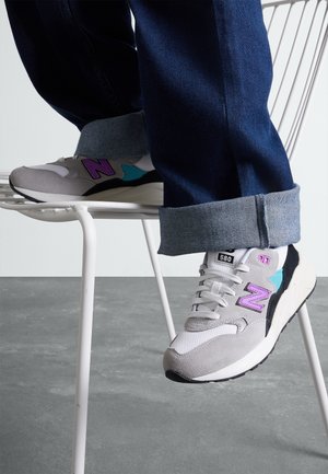 Personne portant des baskets New Balance grises et blanches avec des touches violettes et turquoises, assise sur une chaise en métal blanche avec un jean bleu retroussé.