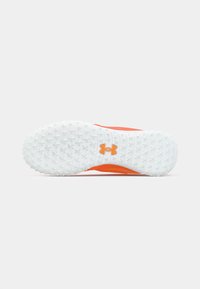 Biała teksturowana podeszwa buta z małymi korkami i pomarańczowym logo Under Armour na środku, umieszczona na jednolitym tle.