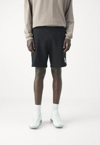 Pantalones cortos deportivos negros con cintura elástica, que cuentan con bolsillos laterales y un parche de logo blanco. Llevados con una camiseta de manga larga beige y zapatillas blancas.