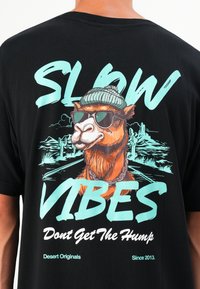 Sort bomulds T-shirt med en farverig illustration af en kamel iført solbriller og en hue, med "SLOW VIBES" tydeligt vist.