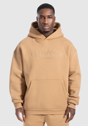 Sudadera oversized de color marrón claro con un bolsillo delantero y puños acanalados. Presenta el logo bordado "SMILODOX ORIGINALS" en el pecho. Textura suave.