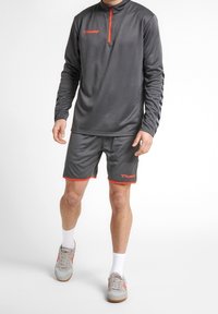 Maglia sportiva grigia a maniche lunghe con zip a un quarto e dettagli arancioni, abbinata a pantaloni corti grigi bordati di arancione, indossata con calzini bianchi e scarpe grigie.