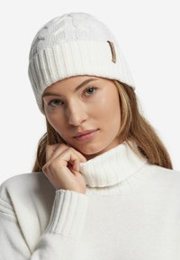 Witte gebreide beanie met geribbelde manchetten, voorzien van een textuur met kabelontwerp en een klein leren labelaccent. Draagt bij een witte turtleneck trui.