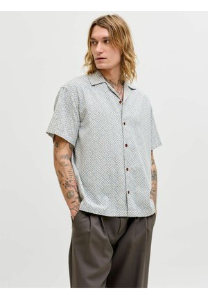 Homme aux cheveux blonds mi-longs et tatouages, portant une chemise à manches courtes à motifs et un pantalon ample foncé, mains dans les poches, regardant vers la gauche.