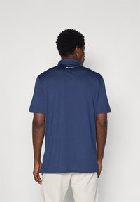 Granatowa koszulka polo z krótkim rękawem, kołnierzykiem, teksturowanym materiałem oraz białym logo Nike na plecach w pobliżu szyi.
