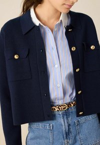 Marine gebreide cropped cardigan met gouden knopenaccenten en twee borstzakken, gedragen over een blauw-wit gestreept shirt. Getextureerde tricot stof.