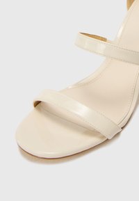Sandal in pelle verniciata beige con due ampie cinghie, punta aperta e una finitura liscia e lucida. Soletta sagomata e tacco leggero.