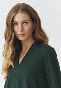 Vêtement vert foncé à col en V avec des manches longues, texture lisse, coutures minimales et coupe décontractée, porté par une personne ayant des cheveux ondulés.