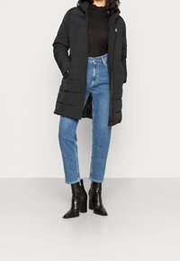 Černá péřová bunda s prošívaným designem, zapínáním na zip a kapucí, kombinovaná se světle modrými džíny s vysokým pasem a černými podpatkovými kotníkovými botami.