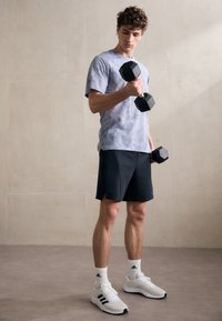 Chemise à manches courtes gris tie-dye, shorts de sport noirs, tenant des haltères noirs, portant des baskets blanches avec des accents noirs, se tenant sur un sol neutre.