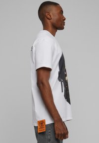 Valge puuvillane T-särk, mille ees on graafiline trükk, oranž ruudukujuline silt küljele, lühikesed varrukad ja lõdvestunud lõige.