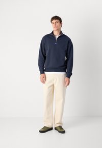 Suéter azul marino con un cuello de cremallera en cuarto, puños acanalados y un acento de logo. Combinado con pantalones color crema y zapatillas verdes.
