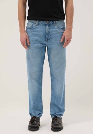 Man draagt lichtblauwe jeans met rechte pijpen, een zwart T-shirt en donkerbruine veterlaarzen, staand tegen een effen achtergrond.