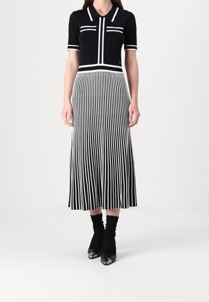 Femme portant un haut noir à manches courtes avec un bord blanc et une jupe midi rayée verticalement noire et blanche ainsi que des talons noirs à bout pointu.