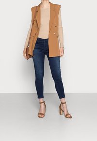 Blazer sin mangas en color tostado, top ajustado de canalé beige, jeans skinny azul oscuro, sandalias de tacón alto marrones con tiras, bolso minimalista.