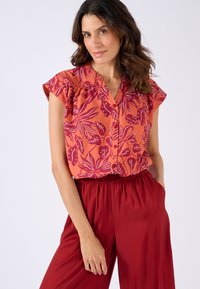 Blusa corta a maniche corte floreale arancione con motivi a foglia viola. Presenta una chiusura a bottoni e dettagli arricciati sulle spalle. Abbinata a pantaloni rossi.