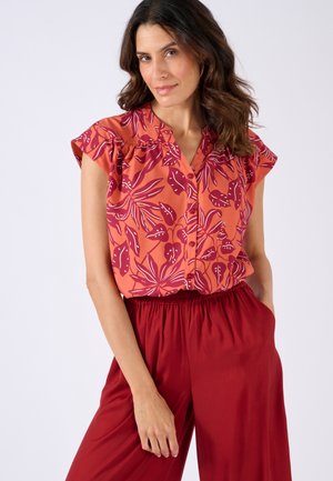 Blusa corta a maniche corte floreale arancione con motivi a foglia viola. Presenta una chiusura a bottoni e dettagli arricciati sulle spalle. Abbinata a pantaloni rossi.