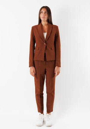 Completo marrone con blazer aderente caratterizzato da un colletto a scatto, chiusura a un bottone e due tasche frontali, abbinato a pantaloni slim-fit e sneakers bianche.