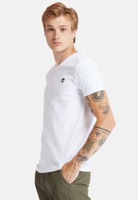 Vit bomulls t-shirt med V-hals som har en liten svart logga. Modellen har tatueringar, solbrun hud och bär olivgröna byxor.