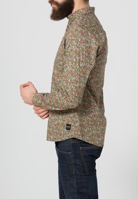 Bloemige blouse met lange mouwen, gekenmerkt door een groene achtergrond met roze en blauwe bloemen, een bandkraag en een merketiket aan de onderrand.