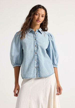 Kvinde med langt bølget hår iført en lyseblå denimskjorte med pufærmer og en flydende hvid nederdel mod en ensfarvet baggrund.